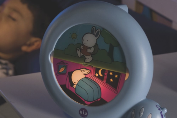 A lighted soft musical or wind noises night light on a shelf, with a sleeping toddler in the background.