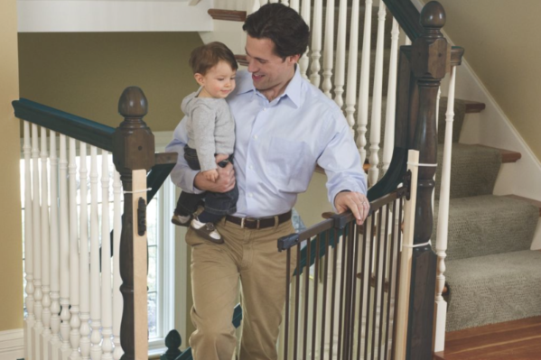 A man holding a baby boy while opening a baby safety gate attached to decorative stair posts, without marring the posts.