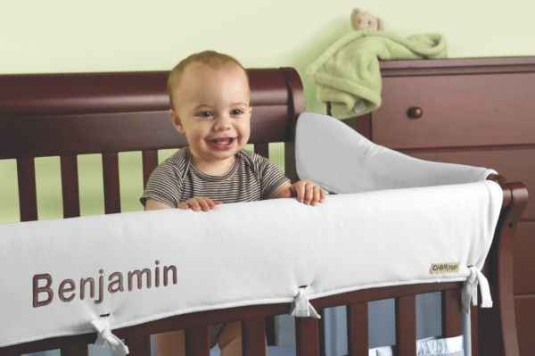 A baby boy standing in his dark wood crib, surrounded by a white personalized, padded top rail protector.