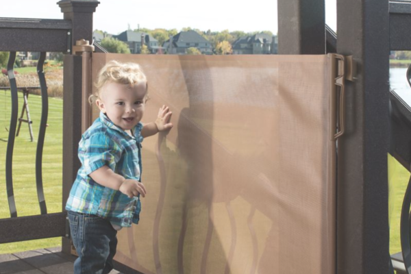 A toddler boy in a blue plaid shirt touching a fabric gate that blocks him from going down outside porch stairs.