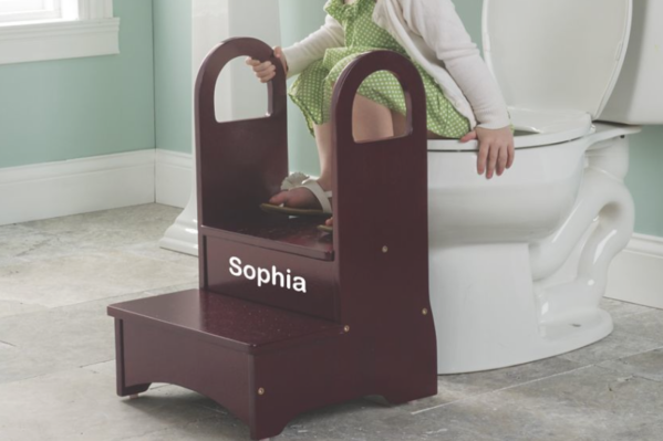 A little girl in a bathroom, using a personalized wooden step stool with safety handles for in front of a full size toilet.