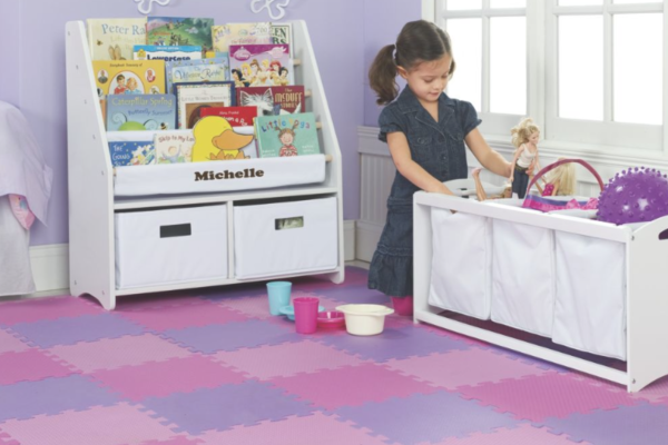 A young girl in her bedroom with a personalized book shelf, a toy bin, and a floor mat of purple and pink locking squares.