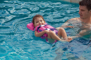 A man holding a floating toddler strapped in a pink safety vest with a head support, enjoying the water in a pool.