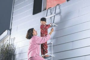 A woman and a boy in pajamas, crawling down a fire escape ladder from an open window to the outside below.