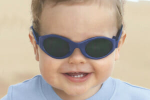 A smiling baby boy at beachside, wearing a light blue top and strap-on blue sunglasses.