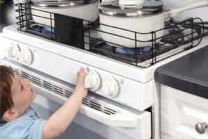 A toddler reaching up to a gas stove top while two pots and the flames below are surrounded by a metal safety gate.