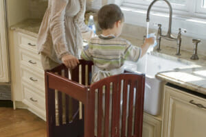 A kitchen scene of a woman and a toddler boy standing on a wooden gated step stool, helping wash his sippy cup.