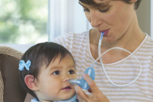 A woman is holding a baby girl in a blue dress on her lap while using a nasal suction to clear the baby's stuffy nose.