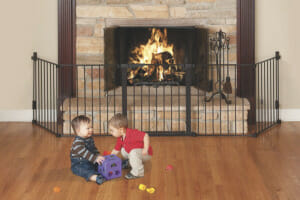 A gated fireplace safety rail attached to the wall, protecting a baby and a toddler playing in front of the fireplace fire.