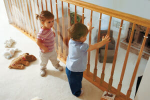 Two babies standing next to a second floor balcony railing that has a plastic guard attached to prevent falling toys.