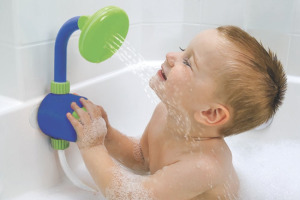 A toddler boy in a bathtub full of bubbles, pushing on a blue and green toy shower head, splashing himself in the face.