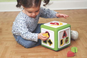 A toddler girl with pigtails, blue top and jeans, playing with a shapes block, trying to put a cross shape in a round hole.