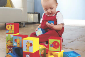 A baby in a red jumpsuit, sitting on the floor, playing with colorful plastic blocks with zoo characters and shapes.