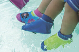 Toddler children's feet clad in colorful pool slippers, splashing the water as they sit on the ledge of a pool.
