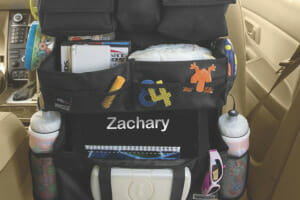 A black organizer with many fabric and mesh pockets, stuffed with baby supplies, is hung on the back of a car's front seat.