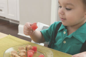 A toddler boy in a teal shirt sitting at a kitchen table, picking up a cube of watermelon from his plate of snack foods.