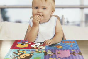 A sitting baby in a bib at a restaurant, enjoying snacks placed on a colorful placemat.