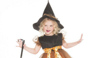A smiling young girl dressed in a fancy orange and black Halloween dress, hat, and striped tights, carrying a fancy broom.