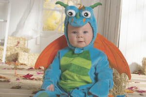 A sitting baby dressed in a blue dragon Halloween costume with green striped belly, orange wings, and dragon head hood.
