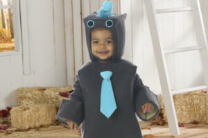A smiling toddler boy in a gray robot Halloween costume with a light blue tie, standing in front of hay bales and leaves.