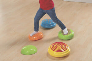 A young girl in a red top and jeans balancing on colorful plastic cushioned floor disks.