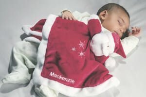 A sleeping baby snuggled with a personalized name on a red and white plush Santa blanket figure.