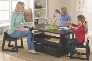 A woman, a boy and a girl sitting at a crafting table with a shelf and large drawer, being creative with scissors and paper.