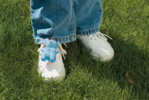 A blue plastic teddy bear child locator, attached to the white shoes of a toddler in jeans who is outside on grass.