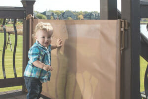 A toddler boy in a blue plaid shirt touching a fabric gate that blocks him from going down outside porch stairs.