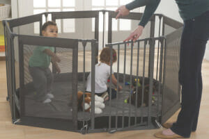 A woman opening the door of a gated mesh play yard, with two toddlers inside, who are playing with toys.
