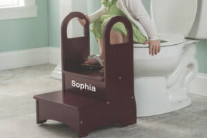 A little girl in a bathroom, using a personalized wooden step stool with safety handles for in front of a full size toilet.