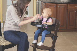 A woman in a kitchen feeding a baby in a dark wooden high chair, with a printed plastic drop sheet on the floor.