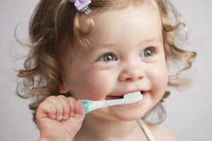 A smiling toddler with a lavender bow in her curly hair, brushing her teeth with a toddler toothbrush.