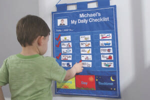 A young boy standing in front of a personalized blue Daily Checklist chart with pictures of daily chores and check marks.