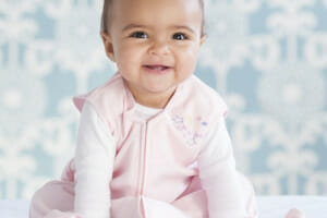 A smiling baby girl wearing a pink full front zip sleep blanket over white jammies, for safety and warmth in the crib.