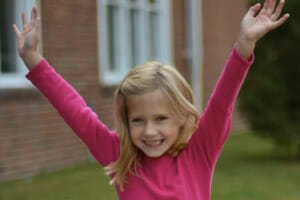 A smiling young girl outside with arms outstretched over her head, wearing a hot pink long sleeve top.