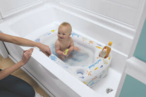 A woman kneeling next to a baby sitting in an air-filled bathtub insert, enjoying the bubbles in the water and a bath toy.