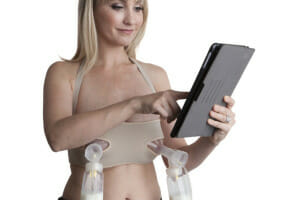 A woman expressing milk automatically with a breast pump and holster while working on a tablet.