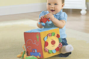 A smiling baby girl wearing a blue top, playing with a colorful soft block set with a hole for the small stuffed toys.