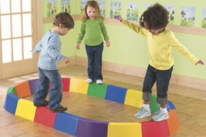 Three young children in a playroom standing and walking on a colorful, circular balance beam.