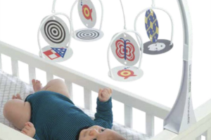 An infant in a crib wearing a blue striped onesie, looking up at a white mobile with colorful simple patterns on disks.