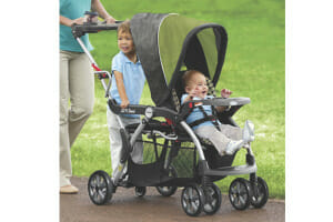 Woman pushing a Graco brand sit and stand stroller with a baby strapped in front and a toddler boy standing in the rear.