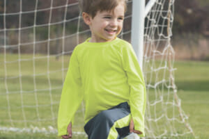 A young boy wearing a lime green long sleeve shirt and gray pants with a foot on a soccer ball, in front of an outside net.