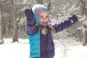 A little girl with outstretched arms playing in the snow in a Cozy Cub purple and turquoise winter jacket and hood.