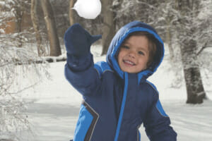 A little boy in a blue Cozy Cub snowsuit, throwing a snowball in front of snow dusted trees and branches.