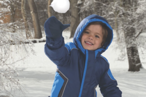 A little boy in a blue Cozy Cub snowsuit, throwing a snowball in front of snow dusted trees and branches.