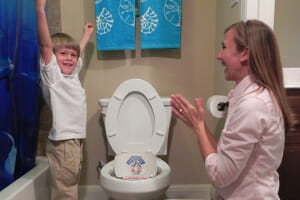 A smiling mom and toddler boy with arms raised in a bathroom, celebrating potty training for boys.
