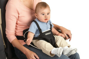 A baby boy in a blue shirt is strapped in a sit seat on a woman's lap, for safe air travel.