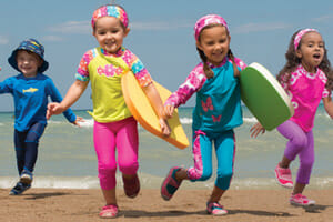 Four young children running on the beach in front of an ocean, wearing colorful Sun Smarties apparel.